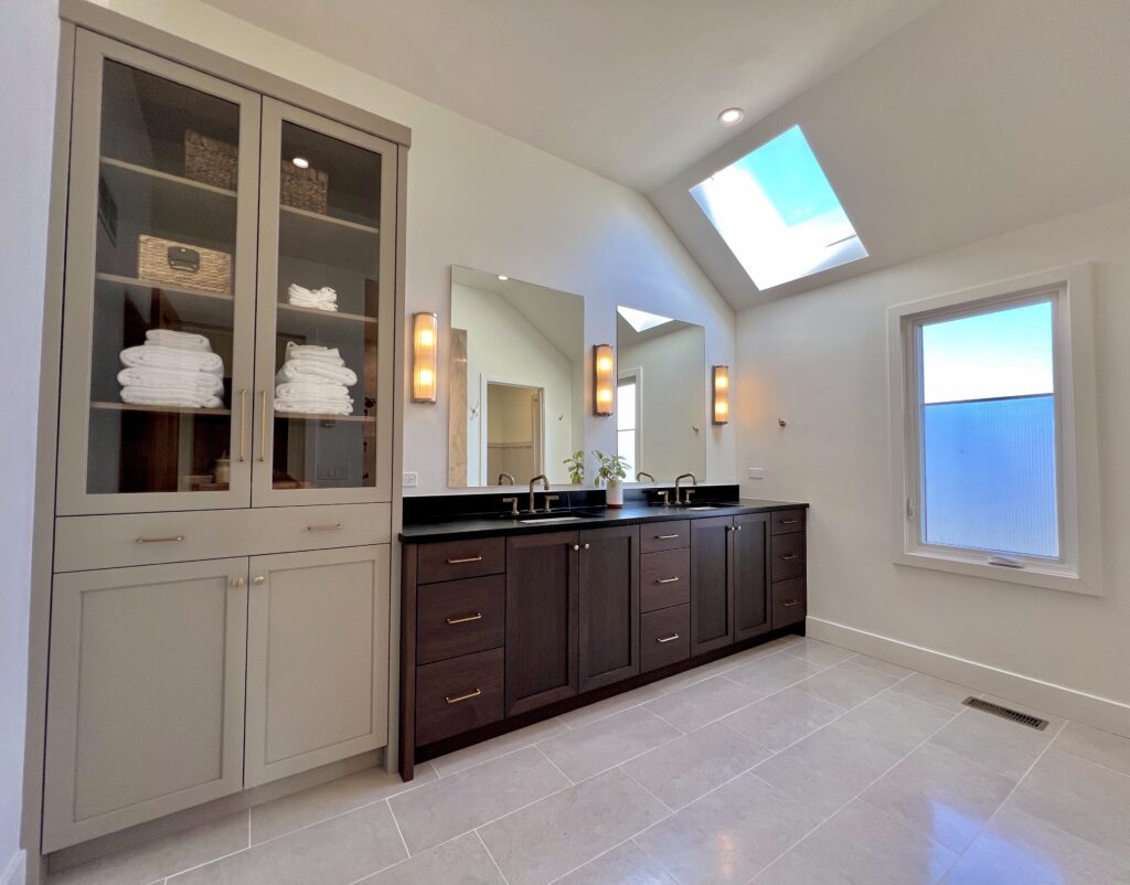 Custom bathroom remodel by Jordan Guide Design featuring a double vanity, built-in storage cabinetry, modern wall sconces, and skylight lighting