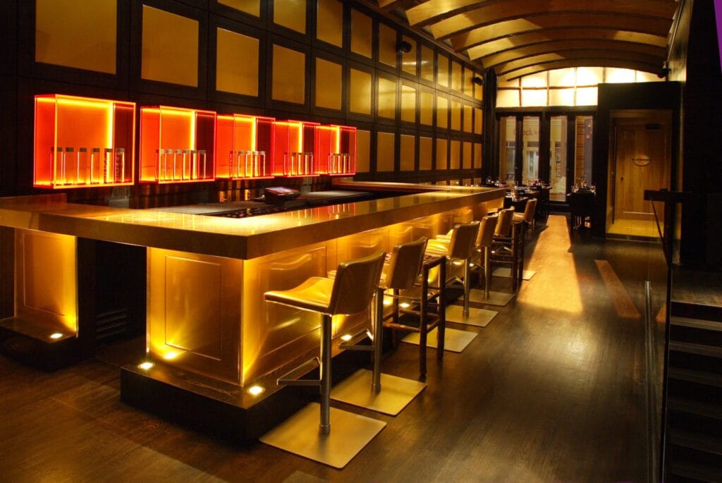 High-end steakhouse bar design featuring illuminated brass bar counter, luxury leather bar seating, dramatic backlit shelving, and modern architectural ceiling lighting