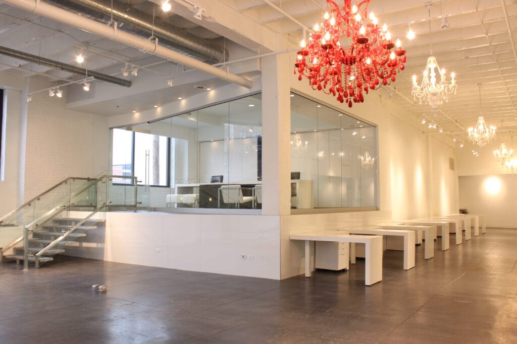 Modern West Loop technology office interior with glass-enclosed executive suite, custom white desks, exposed ceilings, stained concrete floors, and dramatic red chandelier lighting in Chicago.